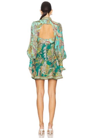 A model shown from the back wearing a short, long-sleeved mini dress with a large open back and a vibrant green and turquoise tropical floral print, paired with beige high-heeled sandals against a white background.