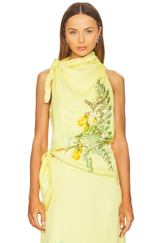 A woman modeling a pale yellow halter dress with a draped wrap-style neckline, side tie, and a botanical print featuring green leaves and yellow flowers on the front, against a plain white background.