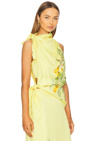 A model wearing a pale yellow sleeveless dress with a high draped collar, side cutouts, a tied waist sash, and a green and yellow floral print on the side, set against a plain white background.