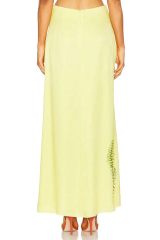 A model from the back wearing a pale yellow maxi skirt with a green fern-like embroidered pattern near the lower right side, paired with brown high-heeled sandals against a white background.
