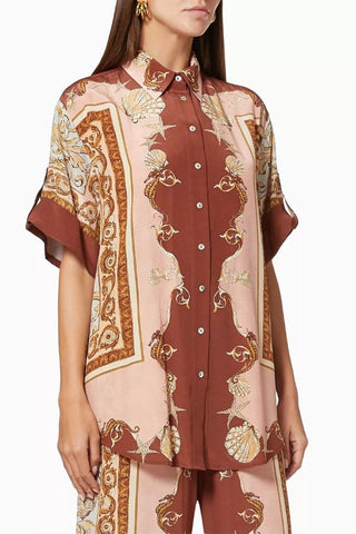 A woman wearing a short-sleeve button-front shirt with a brown and pale pink ornate pattern, paired with matching patterned pants, against a plain light background.