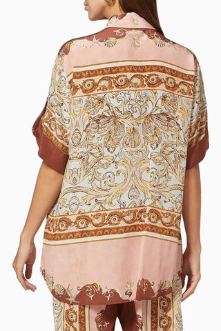 A woman shown from the back wearing a loose short-sleeve top with a dusty pink base and ornate gold and brown baroque-style scrollwork patterns, paired with matching patterned pants against a plain white background.