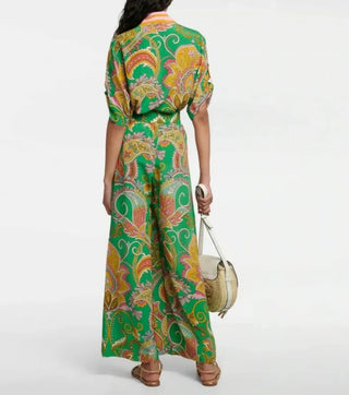 A woman shown from the back wearing a green maxi dress with a colorful paisley pattern in yellow, orange, and pink. She is holding a small beige shoulder bag and wearing brown sandals, set against a plain light gray studio background.