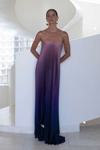 A woman models a strapless pleated maxi dress with a gradient color story, transitioning from a dusty mauve-pink at the sweetheart neckline to a deep purple and finally ending in black at the hem, set against a modern white architectural background.
