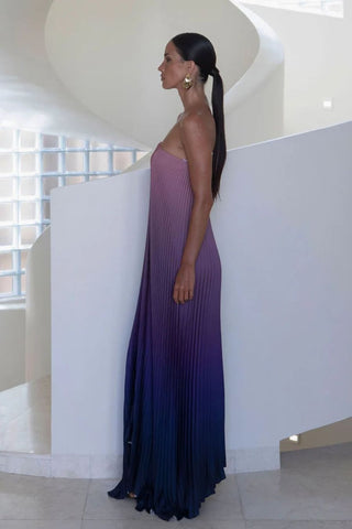 A woman wearing a strapless pleated maxi dress with a vertical ombré gradient from dusty mauve pink to deep purple and navy blue, standing in profile in a modern white architectural setting.