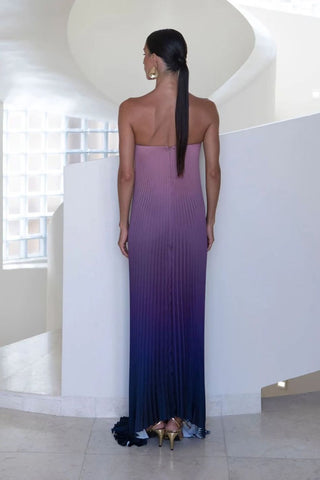A woman stands with her back to the camera wearing a strapless pleated maxi dress with a vertical ombre gradient from dusty lavender-purple at the shoulders to deep navy blue at the hem, in a modern white architectural setting.