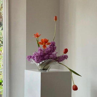 Clear glass vase with red tulips and purple lilacs on a white pedestal against a neutral wall.