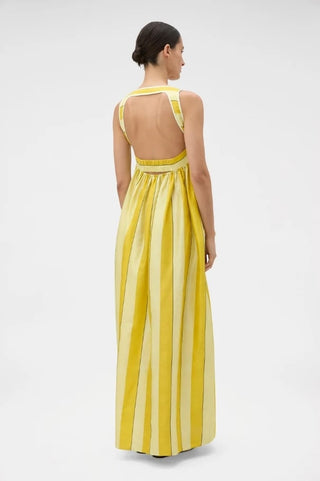 A woman shown from the back wearing a sleeveless yellow and white vertically striped maxi dress with a low open back, photographed against a plain light gray studio background.