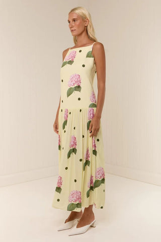 A woman modeling a sleeveless cream maxi dress with a pink and green floral print and black polka dots, paired with white pointed-toe heels, in a minimalist cream-colored studio setting.