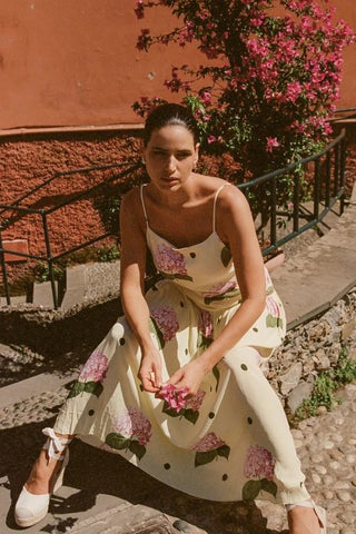 Woman in a floral dress sitting on a stone path with pink flowers and a red wall in the background
