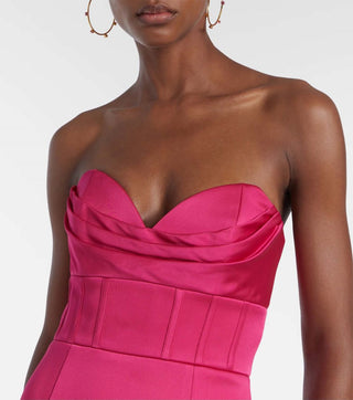 A close-up of a model wearing a bright fuchsia pink strapless satin dress with a sweetheart neckline, ruched bodice, and structured waist, paired with gold hoop earrings against a light gray background.