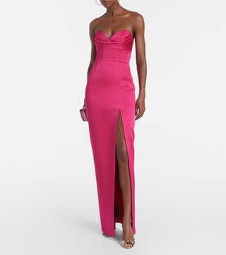 A woman models a bright fuchsia pink strapless gown with a sweetheart neckline, structured bodice, and a high thigh slit, paired with metallic sandals against a light gray studio background.