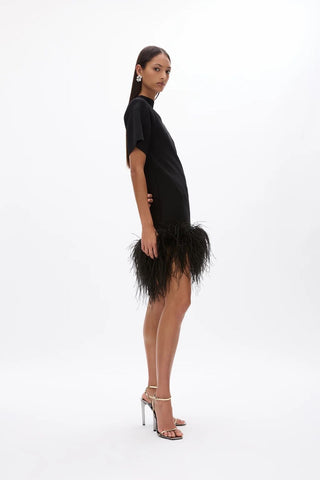 A model wearing a black short-sleeve mini dress with a feathered hem, paired with metallic high-heeled sandals, photographed against a seamless white studio background.