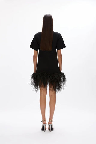 A model viewed from behind wearing a black short-sleeve mini dress with a feathered hem, paired with black high-heeled sandals, against a plain white studio background.