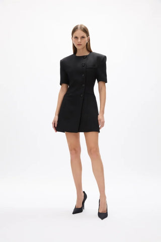 A model wearing a black short-sleeve mini dress with structured shoulders and a button front, paired with black pointed-toe heels, against a plain white studio background.
