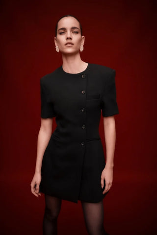 A model wearing a black short-sleeve mini dress with structured shoulders and a button-front detail, posed against a solid deep red background.