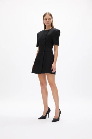 A woman modeling a black short-sleeve mini dress with structured shoulders and a button front, paired with black pointed-toe heels, against a plain white studio background.