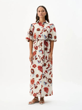 A woman stands against a plain white studio background wearing a long-sleeve collared maxi dress with a cream base and a red, brown, and blue floral print. She is wearing brown strappy sandals.