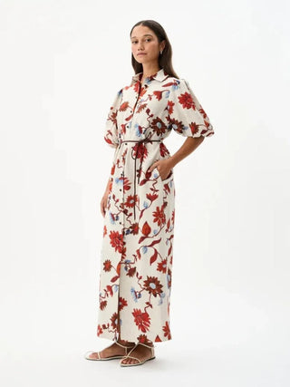 A woman modeling a cream-colored maxi shirt dress with a red and blue floral print, featuring short puff sleeves, a button-up front, and a matching tie belt, set against a plain white studio background.
