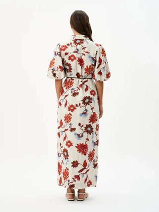 A woman viewed from the back wearing a cream-colored maxi dress with a red, blue, and brown floral print, styled with brown heeled sandals against a plain white studio background.
