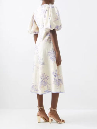 A woman shown from the back wearing a cream midi dress with a light purple palm tree print, puff sleeves, and a gathered waist, paired with beige strappy high-heeled sandals against a plain white studio background.